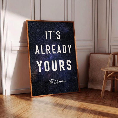 A framed motivational art print leaning against a white paneled wall on a wooden floor. The print features the quote 'IT'S ALREADY YOURS - To The Universe' in white text over a dark, starry space background, enclosed in a light brown wood frame.