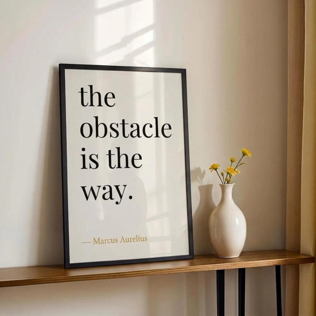 Framed quote 'the obstacle is the way.' by Marcus Aurelius on a wall with a vase of flowers below.