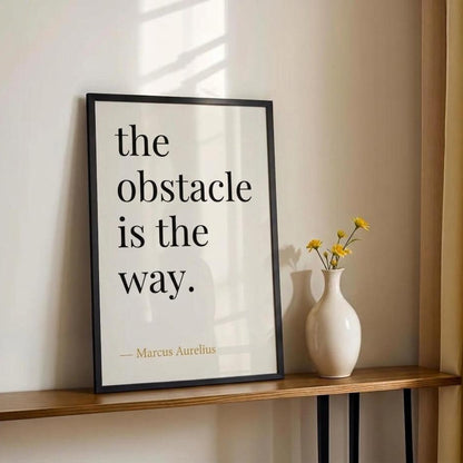 Framed quote 'the obstacle is the way.' by Marcus Aurelius on a wall with a vase of flowers below.