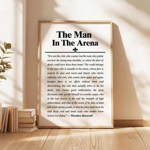 A framed motivational poster titled 'The Man In The Arena' with a quote attributed to Theodore Roosevelt, displayed on a wooden surface next to books and a plant.