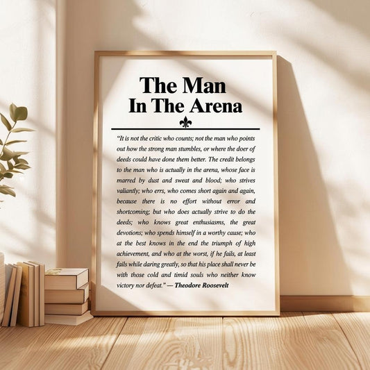 A framed motivational poster titled 'The Man In The Arena' with a quote attributed to Theodore Roosevelt, displayed on a wooden surface next to books and a plant.