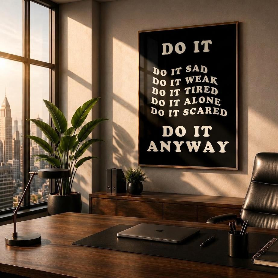 A framed motivational wall art print with the text 'DO IT ANYWAY' in white serif lettering on a black background, hanging in a modern office setting above a wooden desk with a black leather chair.