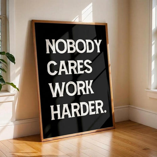 A framed motivational wall art print leaning against a beige wall in a sunlit room with wood floors and windows. The print has a black background with large white text reading 'NOBODY CARES WORK HARDER.