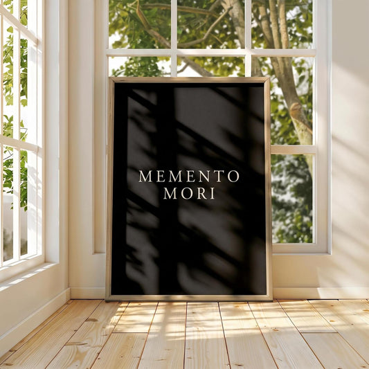 A vertical black art print with the words 'MEMENTO MORI' in white serif lettering, leaning against a white wall in a sunlit room with wood floors and large windows.