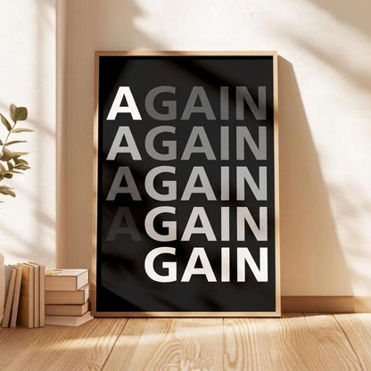 A framed typographic wall art print leaning against a beige wall on a wooden floor, with stacked books and a plant nearby. The print has a black background, large repeated text reading 'AGAIN AGAIN AGAIN AGAIN GAIN' in white and gray, and a light wood frame.