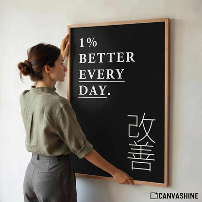 A woman in a sage green shirt and gray trousers stands beside a framed blackboard with the motivational phrase '1% BETTER EVERY DAY.' in white text and large white Japanese characters, against a plain light wall.