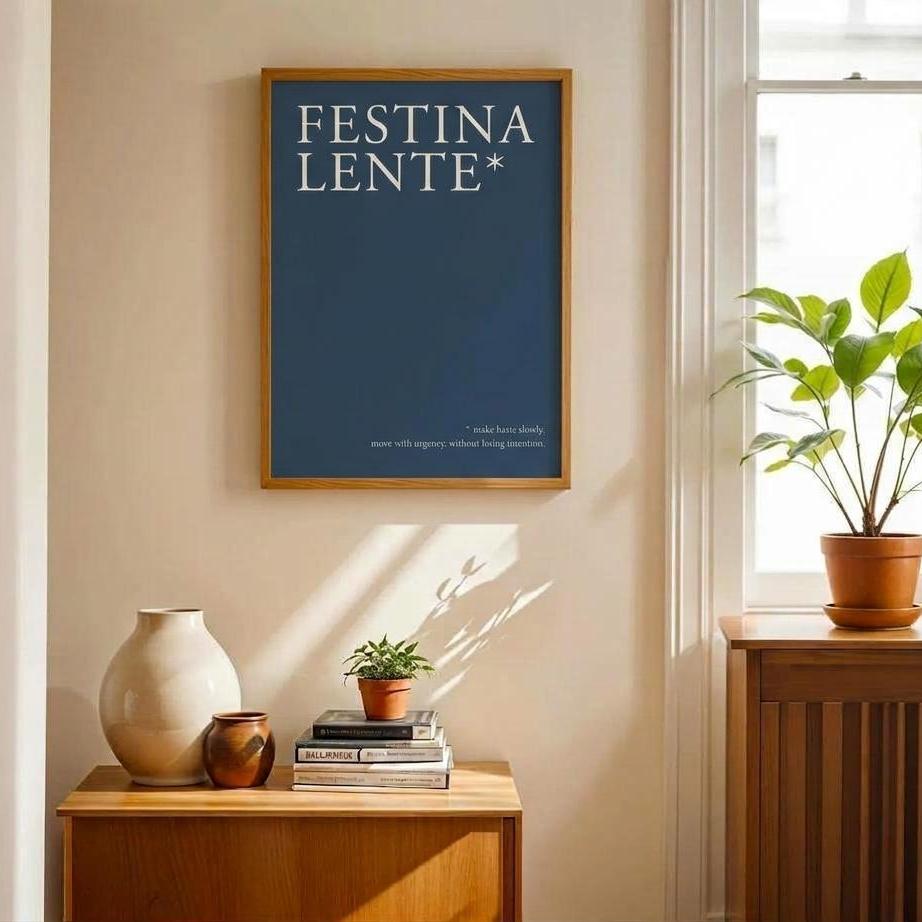 A framed art print featuring the text 'FESTINA LENTE*' in white serif lettering on a dark blue background, hanging on a beige wall in a sunlit room with a wooden cabinet, books, and a potted plant.