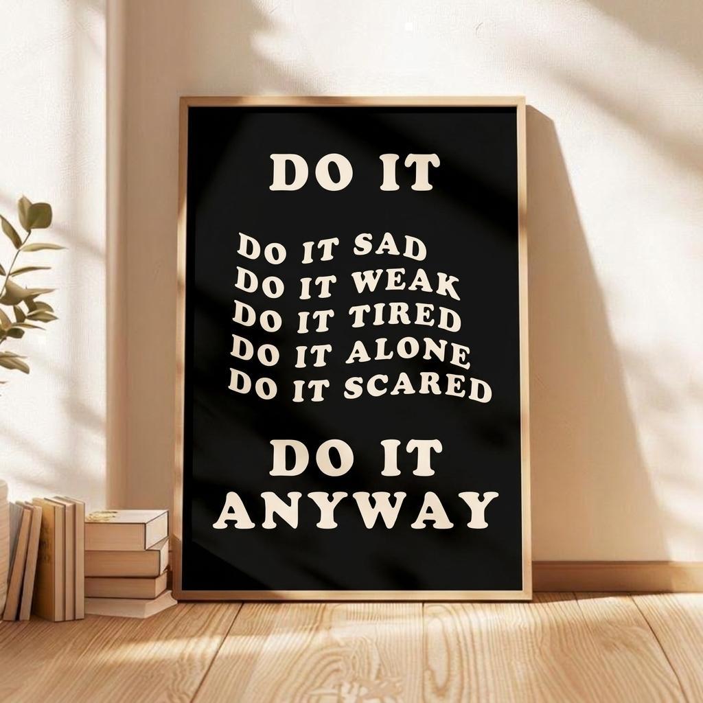 A framed motivational poster with the text 'DO IT ANYWAY' in cream lettering on a black background, leaning against a beige wall on a wooden floor next to stacked books and a plant.