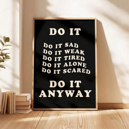 A framed motivational poster with the text 'DO IT ANYWAY' in cream lettering on a black background, leaning against a beige wall on a wooden floor next to stacked books and a plant.