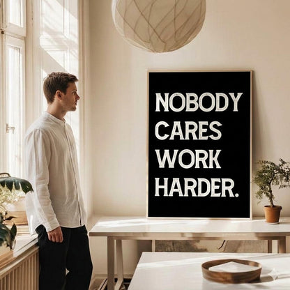 A man in a white shirt and black pants stands in a bright room looking at a framed poster that reads 'NOBODY CARES WORK HARDER.' in bold white text on a black background.