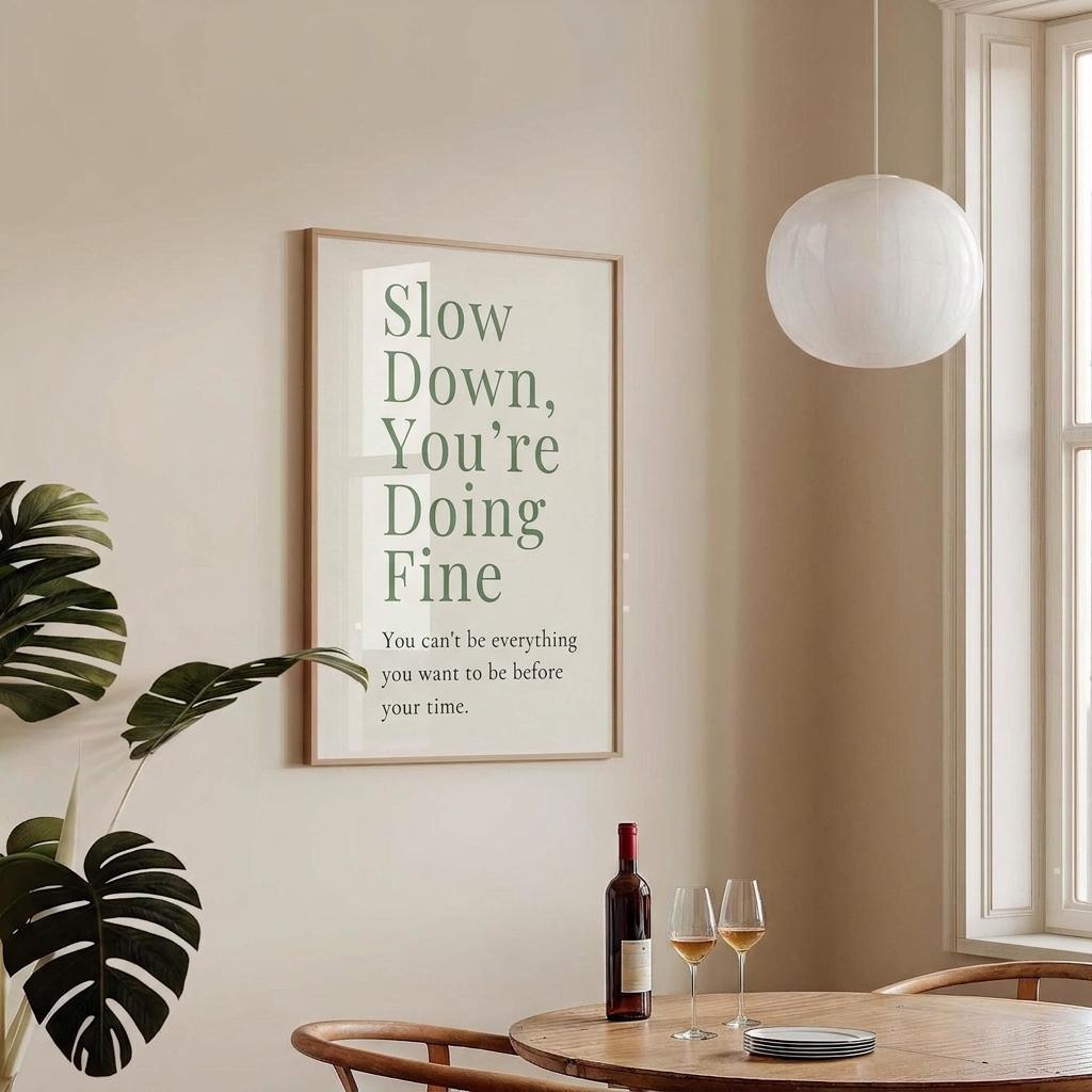 A framed wall art print with the phrase 'Slow Down, You're Doing Fine' in green text on a cream background, hanging on a beige wall in a styled dining room with a table, wine bottle, two wine glasses, a hanging pendant light, and a window.