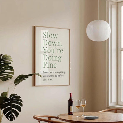 A framed wall art print with the phrase 'Slow Down, You're Doing Fine' in green text on a cream background, hanging on a beige wall in a styled dining room with a table, wine bottle, two wine glasses, a hanging pendant light, and a window.
