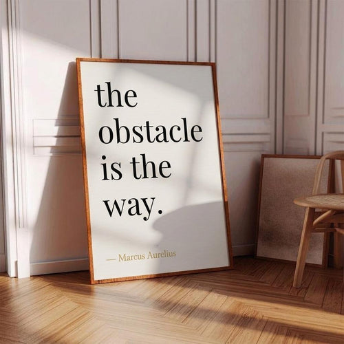 Framed poster with 'the obstacle is the way' quote by Marcus Aurelius in a room with wooden floor and chair.