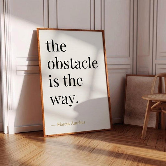 Framed poster with 'the obstacle is the way' quote by Marcus Aurelius in a room with wooden floor and chair.