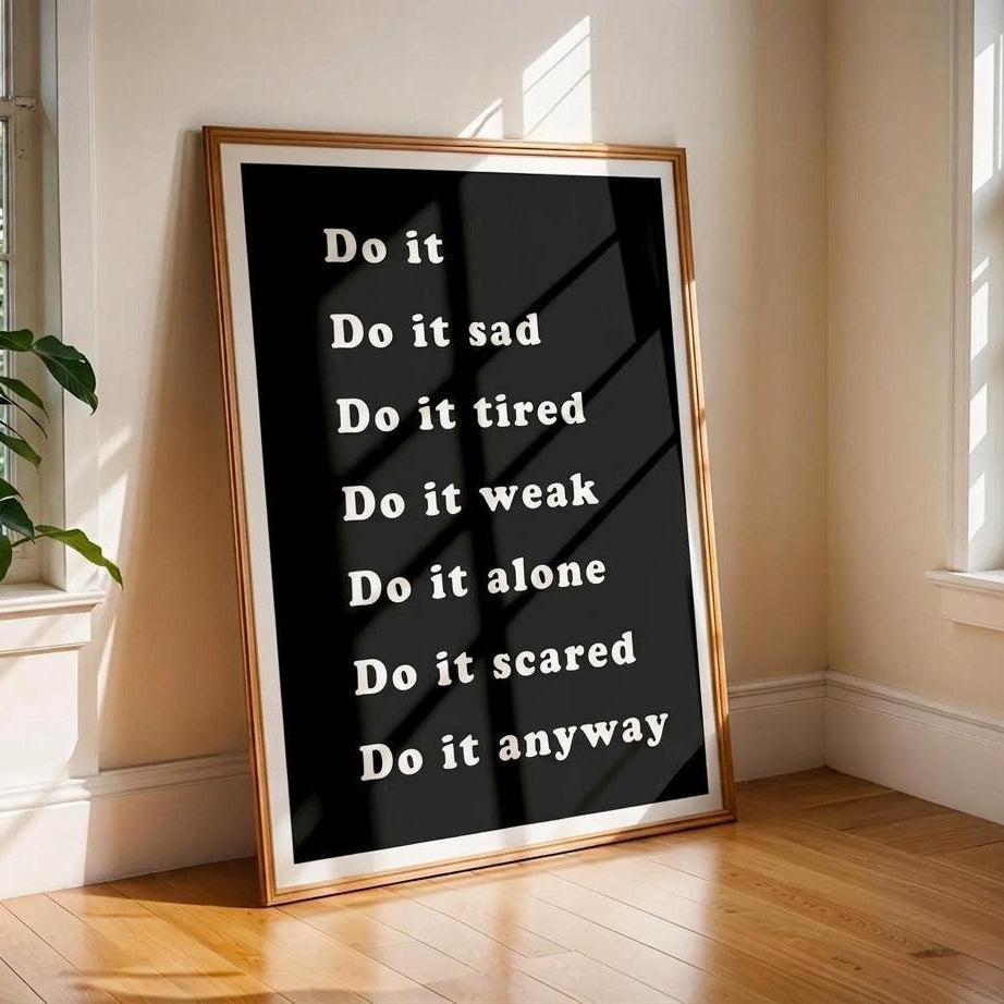 A framed motivational poster with the text 'Do it sad Do it tired Do it weak Do it alone Do it scared Do it anyway' in white on a black background, leaning against a beige wall on a wooden floor in a sunlit room.