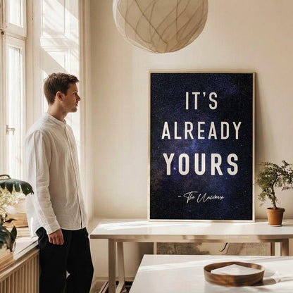 A man in a white shirt and black pants stands in a bright room looking at a framed poster on the wall. The poster has a dark blue galaxy background with the text 'IT'S ALREADY YOURS - The Universe'