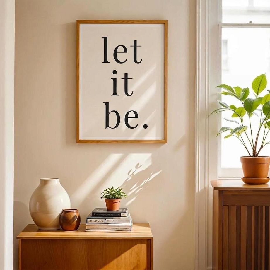 A framed wall art print with the text 'let it be.' in black serif lowercase letters on a white background, hanging in a warm beige room above a wooden sideboard with decorative vases, books, and a small plant.