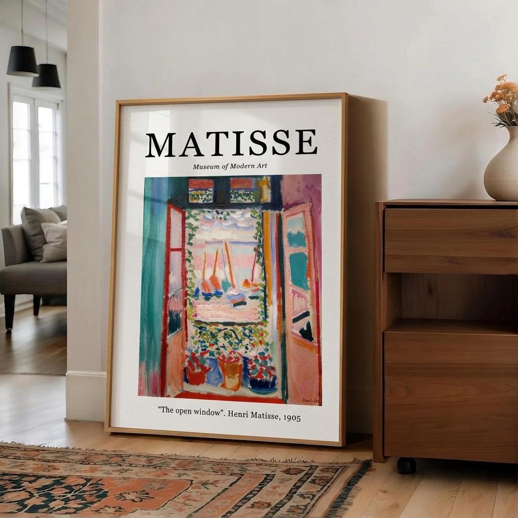 A framed art print of Henri Matisse's 'The open window' leaning against a white wall in a modern living room setting next to a wooden cabinet.