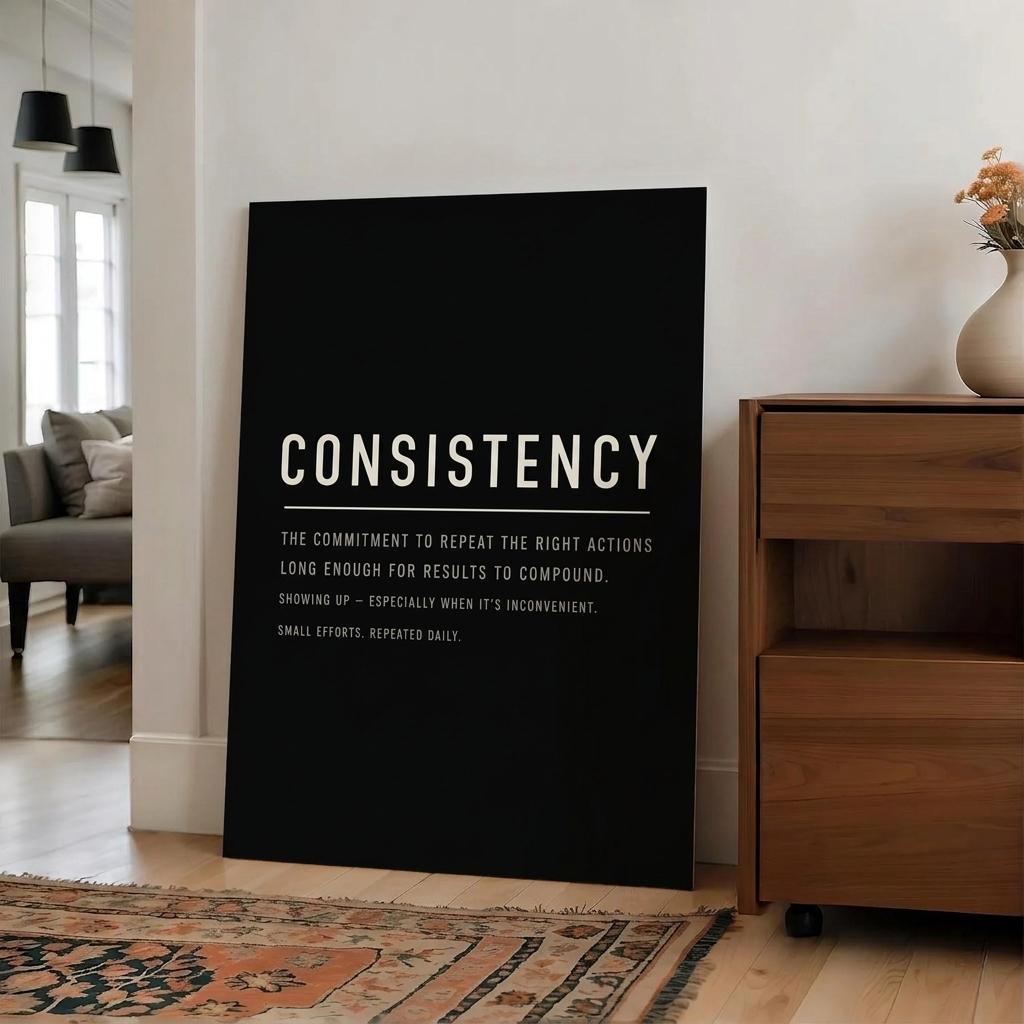 A large black rectangular wall art print leaning against a white wall in a modern living room. The print features the word 'CONSISTENCY' in bold white uppercase letters, with smaller white text underneath defining consistency.