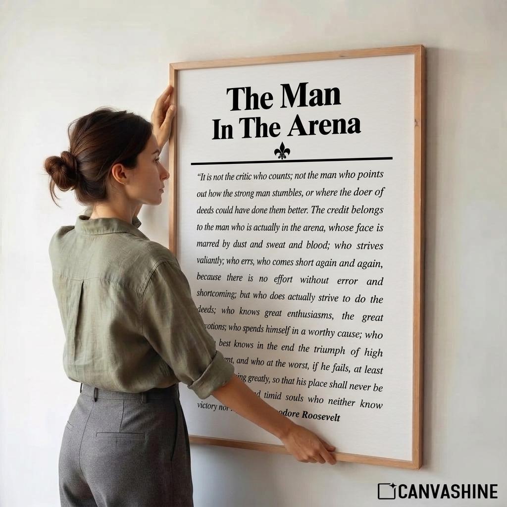 A woman in a sage green shirt and gray pants is holding a large framed motivational poster on a white wall. The poster features the text 'The Man In The Arena' with a quote attributed to Roosevelt.