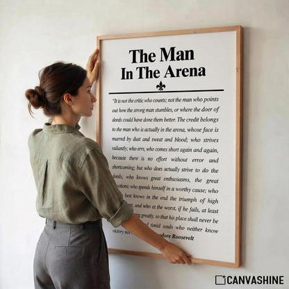 A woman in a sage green shirt and gray pants is holding a large framed motivational poster on a white wall. The poster features the text 'The Man In The Arena' with a quote attributed to Roosevelt.