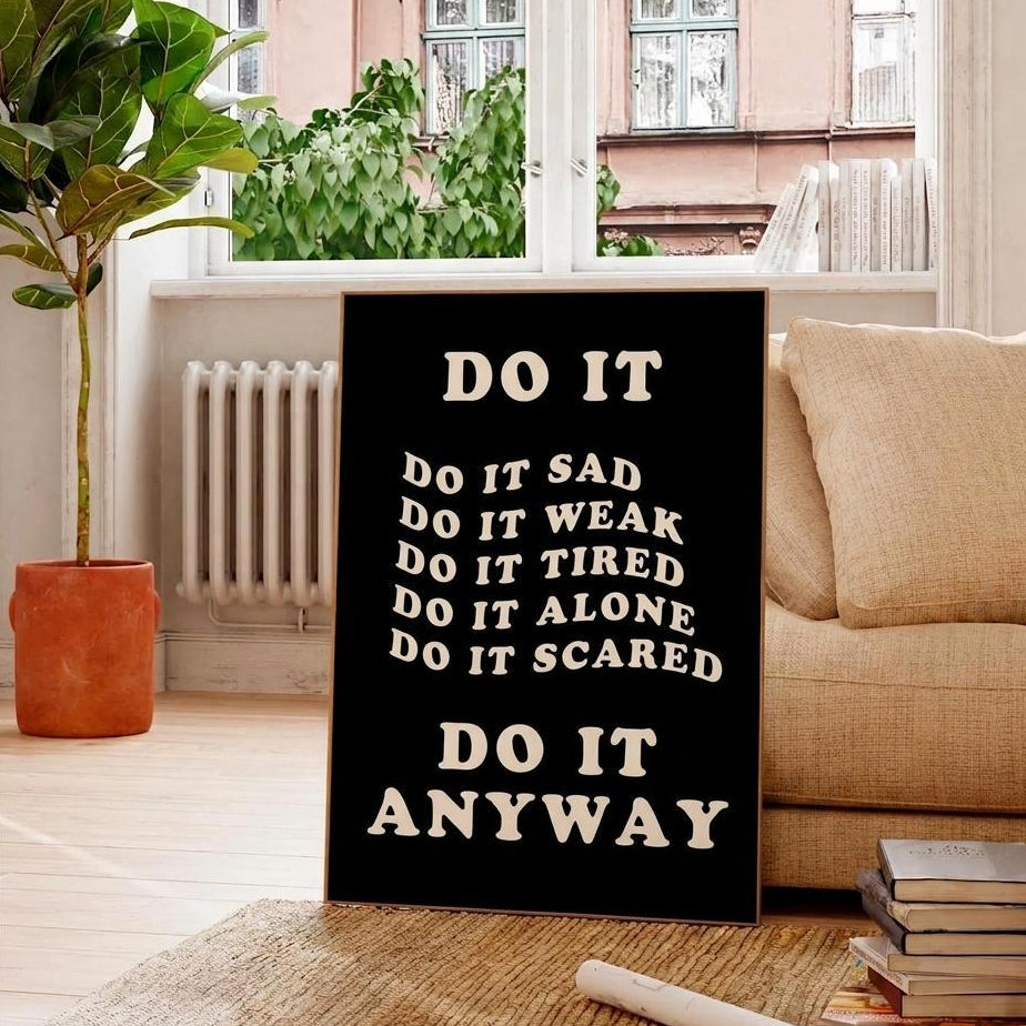 A black rectangular wall sign with white text reading 'DO IT ANYWAY' and motivational phrases in the middle, displayed in a living room setting next to a beige sofa and a potted plant.