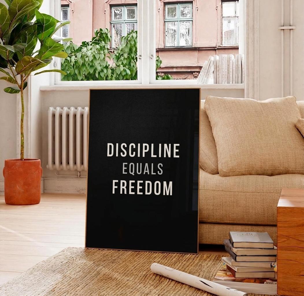 A black framed wall art print with the white text 'DISCIPLINE EQUALS FREEDOM' leaning against a beige sofa in a bright living room with a window, plant, and books.