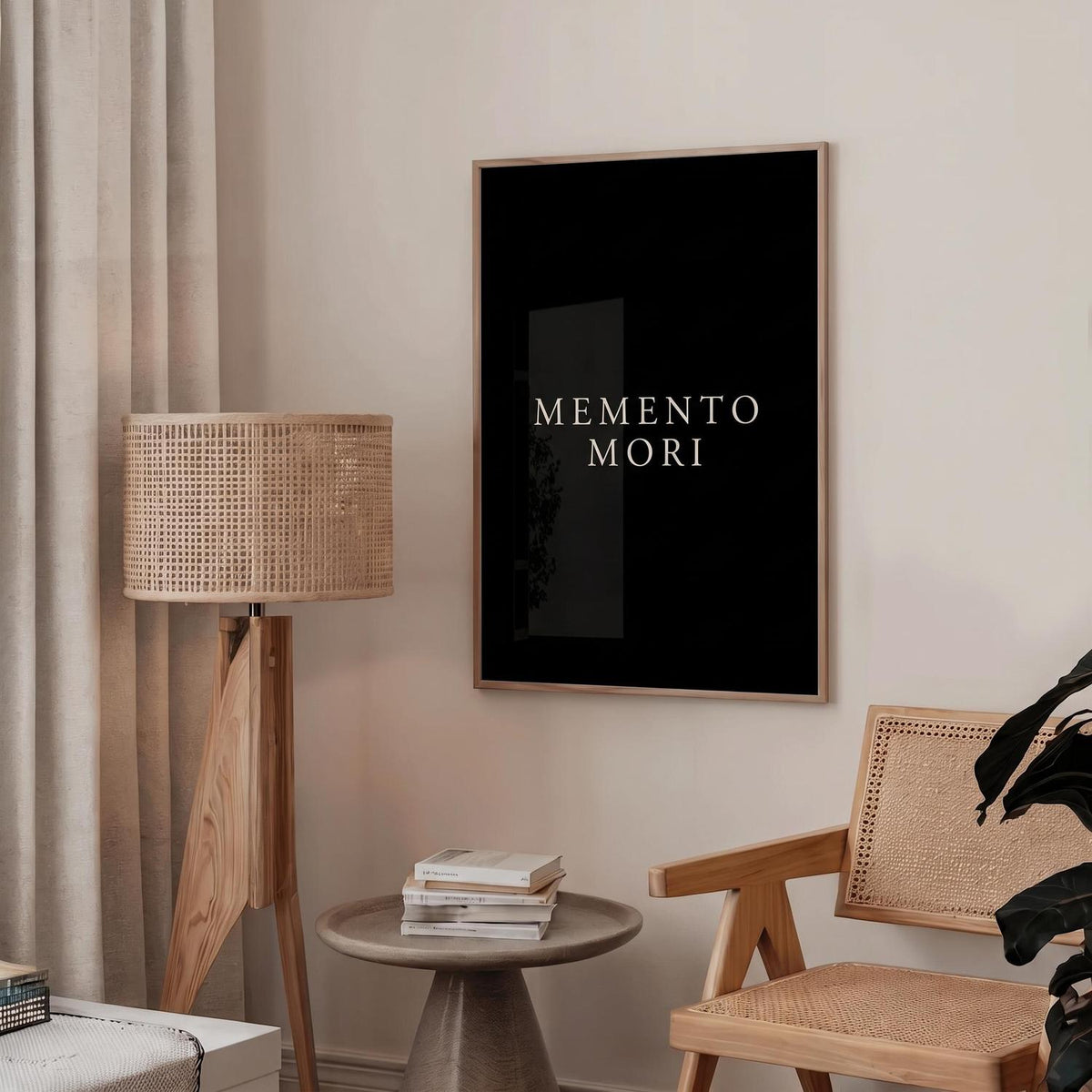 A framed black wall art print with the words 'MEMENTO MORI' in white serif text hangs on a beige wall in a styled room with a floor lamp, side table, chair, and plant.