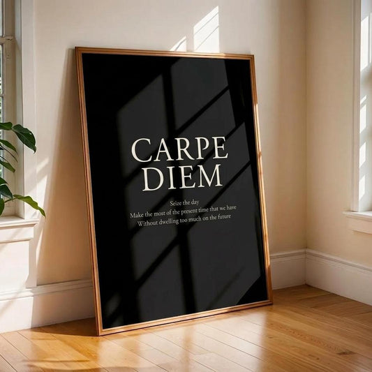 A framed black art print leaning against a beige wall on a wooden floor in a sunlit room. The print has a thin gold frame and white text reading 'CARPE DIEM' with smaller quote text below.