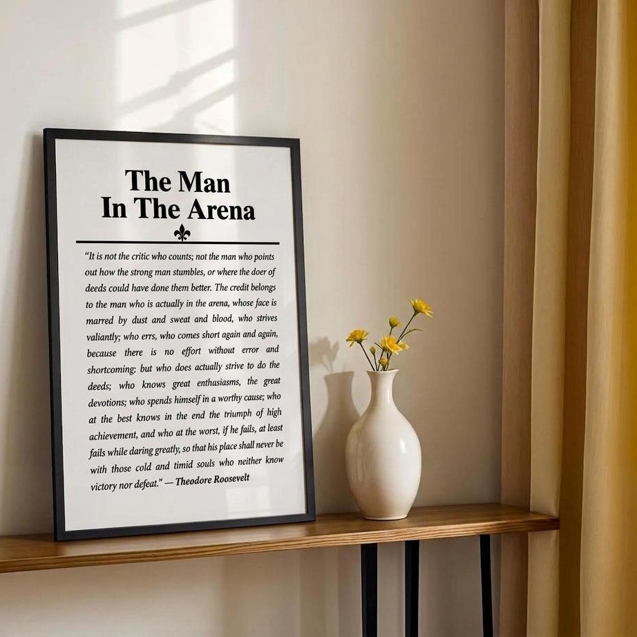A framed black-and-white motivational poster titled "The Man In The Arena" leaning on a wooden shelf next to a small vase with yellow flowers against a beige wall.