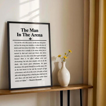 A framed black-and-white motivational poster titled "The Man In The Arena" leaning on a wooden shelf next to a small vase with yellow flowers against a beige wall.