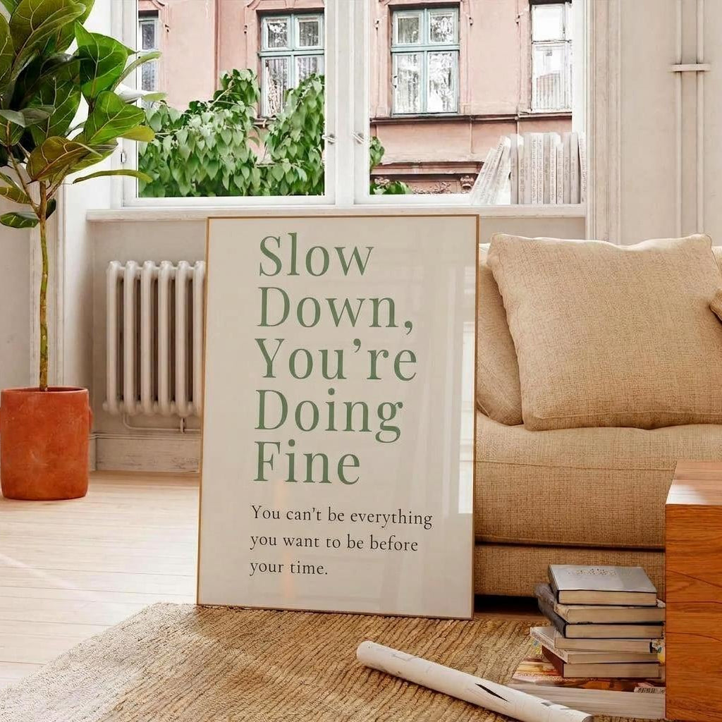 A framed typographic wall art print leaning against a beige sofa in a bright living room. The print reads, 'Slow Down, You're Doing Fine' with smaller text below, 'You can't be everything you want to be before your time.