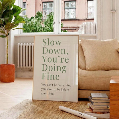 A framed typographic wall art print leaning against a beige sofa in a bright living room. The print reads, 'Slow Down, You're Doing Fine' with smaller text below, 'You can't be everything you want to be before your time.