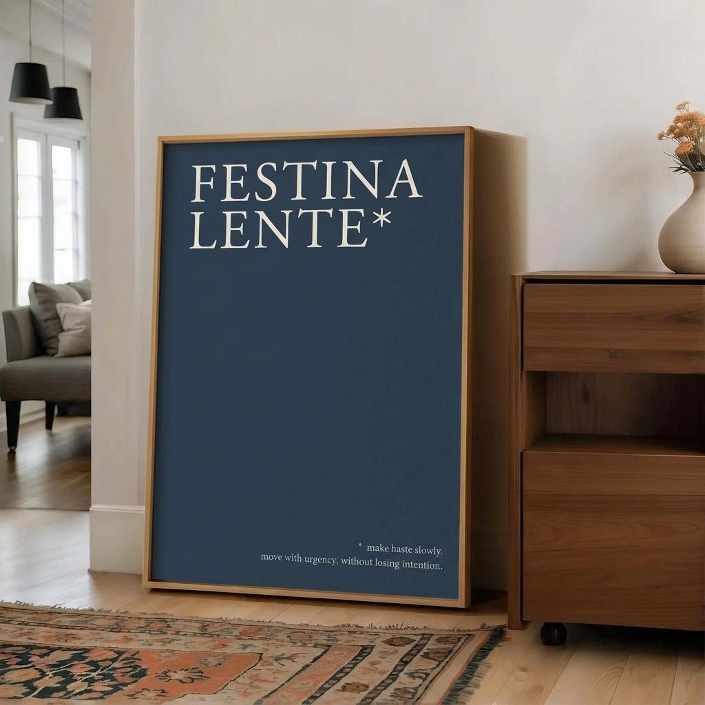 A framed art print featuring the text 'FESTINA LENTE*' in white on a dark blue background, leaning against a white wall in a modern living room setting next to a wooden cabinet.