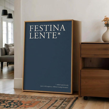 A framed art print featuring the text 'FESTINA LENTE*' in white on a dark blue background, leaning against a white wall in a modern living room setting next to a wooden cabinet.