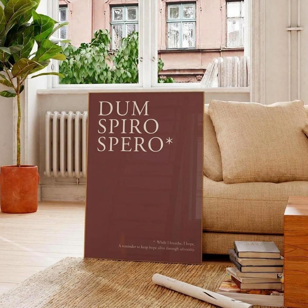 A tall, slim book or art print with the title 'DUM SPIRO SPERO*' in white serif text on a dark reddish-brown cover, standing on a woven rug in a bright living room beside a beige sofa and a potted plant.