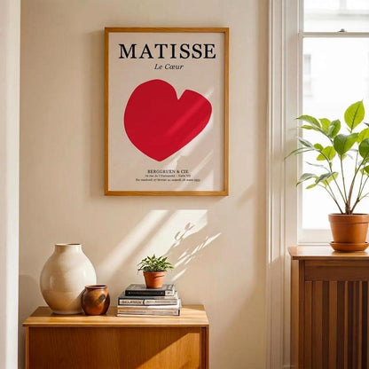 A framed Matisse 'Le Coeur' poster with a large red heart hangs on a beige wall in a sunlit room. Below the poster is a wooden sideboard with a ceramic vase, small books, and a potted plant, while a larger plant sits by the window on a matching cabinet.