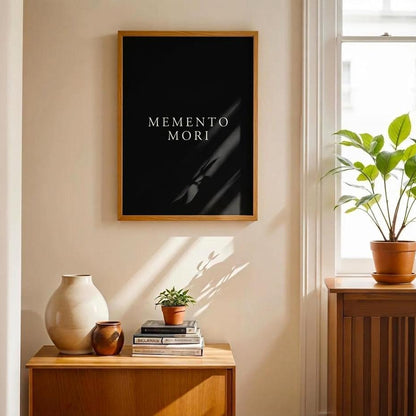 A framed black art print with the text 'MEMENTO MORI' in white serif letters, hanging on a beige wall in a sunlit room with a wooden side table, ceramic vases, and a potted plant by the window.