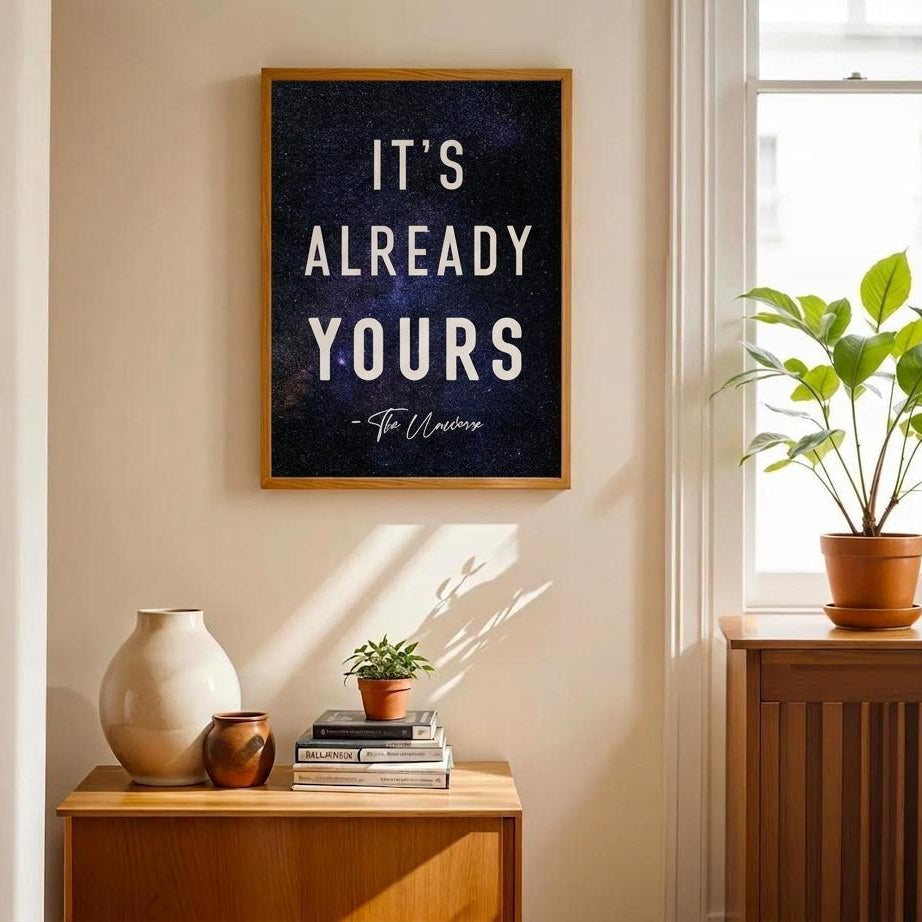 A framed wall art print with the text 'IT'S ALREADY YOURS' hangs on a beige wall in a sunlit room. The print has a dark textured background and a light wood frame.