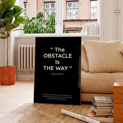 A black vertical wall art print leaning against a beige sofa in a bright living room. The print features a white quote by Marcus Aurelius: "The OBSTACLE IS THE WAY".
