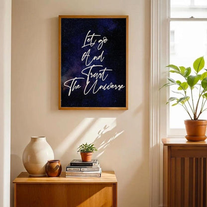 A framed wall art print hangs on a beige wall in a sunlit room. The print has a dark navy and black starry background with white script text reading, 'Let go and forget the universe.