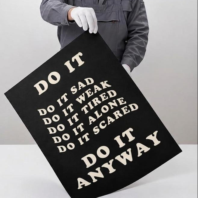 A person in a gray work jacket and white gloves holds a large black canvas print with white text reading 'DO IT ANYWAY' against a light gray studio background. The left side features a beige panel with text describing professional giclée quality and product features.