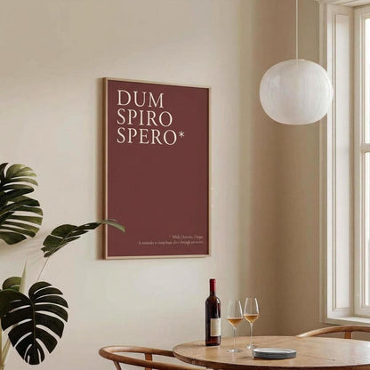A framed wall art print with the text 'DUM SPIRO SPERO*' in white on a muted burgundy background, hanging on a beige wall in a bright dining room with a table, wine bottle, two wine glasses, a hanging pendant light, and a window.