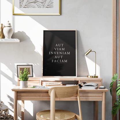 A framed black art print with the Latin text 'AMOR FATI CARPE DIEM' in white serif lettering, displayed on a light wood desk in a modern, sunlit home office setting.