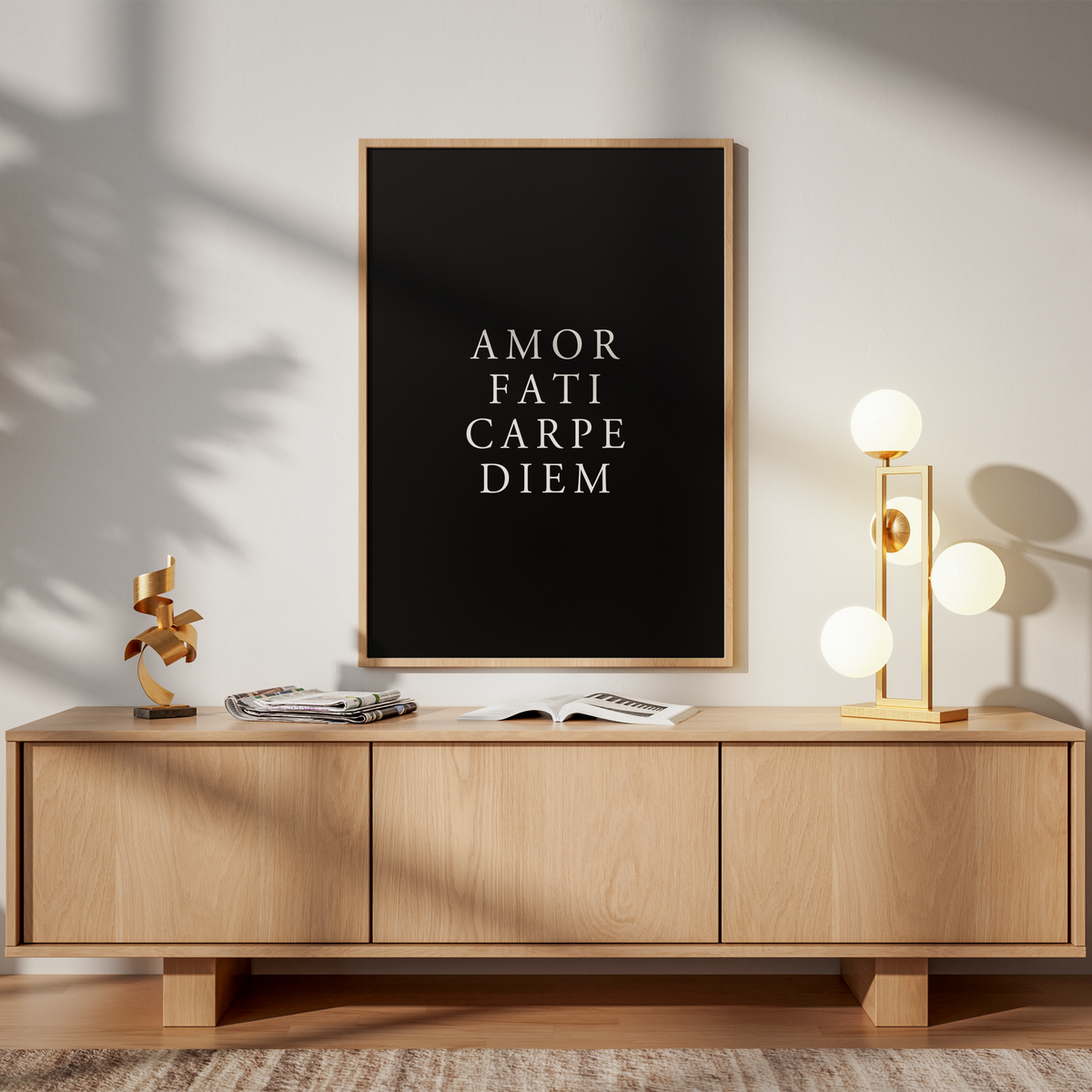A framed black wall art print with white serif text reading 'AMOR FATI CARPE DIEM' hangs above a light wood sideboard in a styled interior. The room has warm sunlight and shadows, with a gold floor lamp, books, and a chair visible.