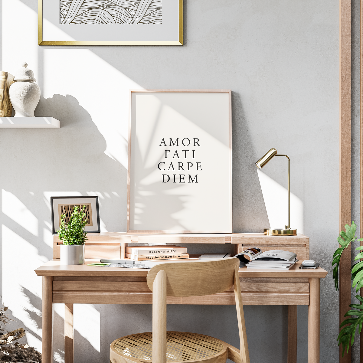 A framed art print with the Latin text 'AMOR FATI CARPE DIEM' in black serif font on a cream background, styled in a warm, modern home office setting with a wooden desk, chair, lamp, and plants.