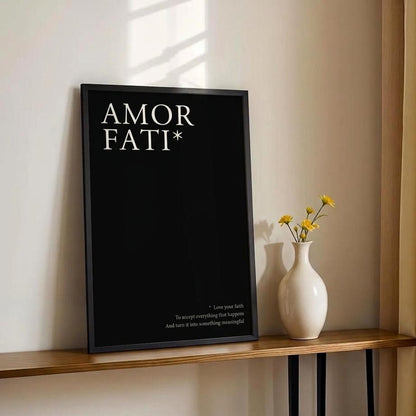 A black framed poster with the text 'AMOR FATI*' in white, leaning on a wooden console table next to a white vase with yellow flowers against a beige wall with warm sunlight and curtains.