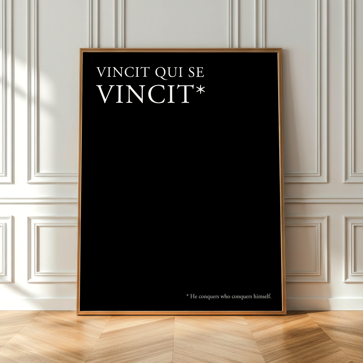 A vertical black poster with a thin gold frame leans against a white paneled wall on a light wood herringbone floor. The poster features the white text 'VINCIT QUI SE VINCIT*' at the top and a small footnote at the bottom reading '*He compares who compares himself.