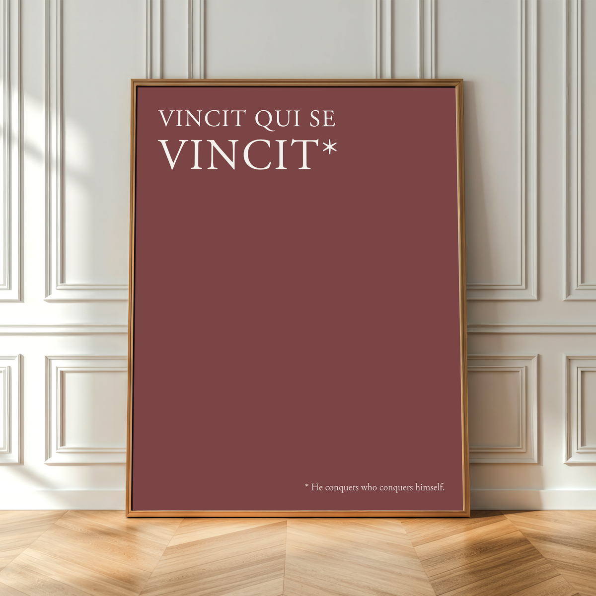 A framed minimalist art print with a terracotta red background and white text reading 'VINCIT QUI SE VINCIT*' leaning against a white paneled wall on a light wood herringbone floor.