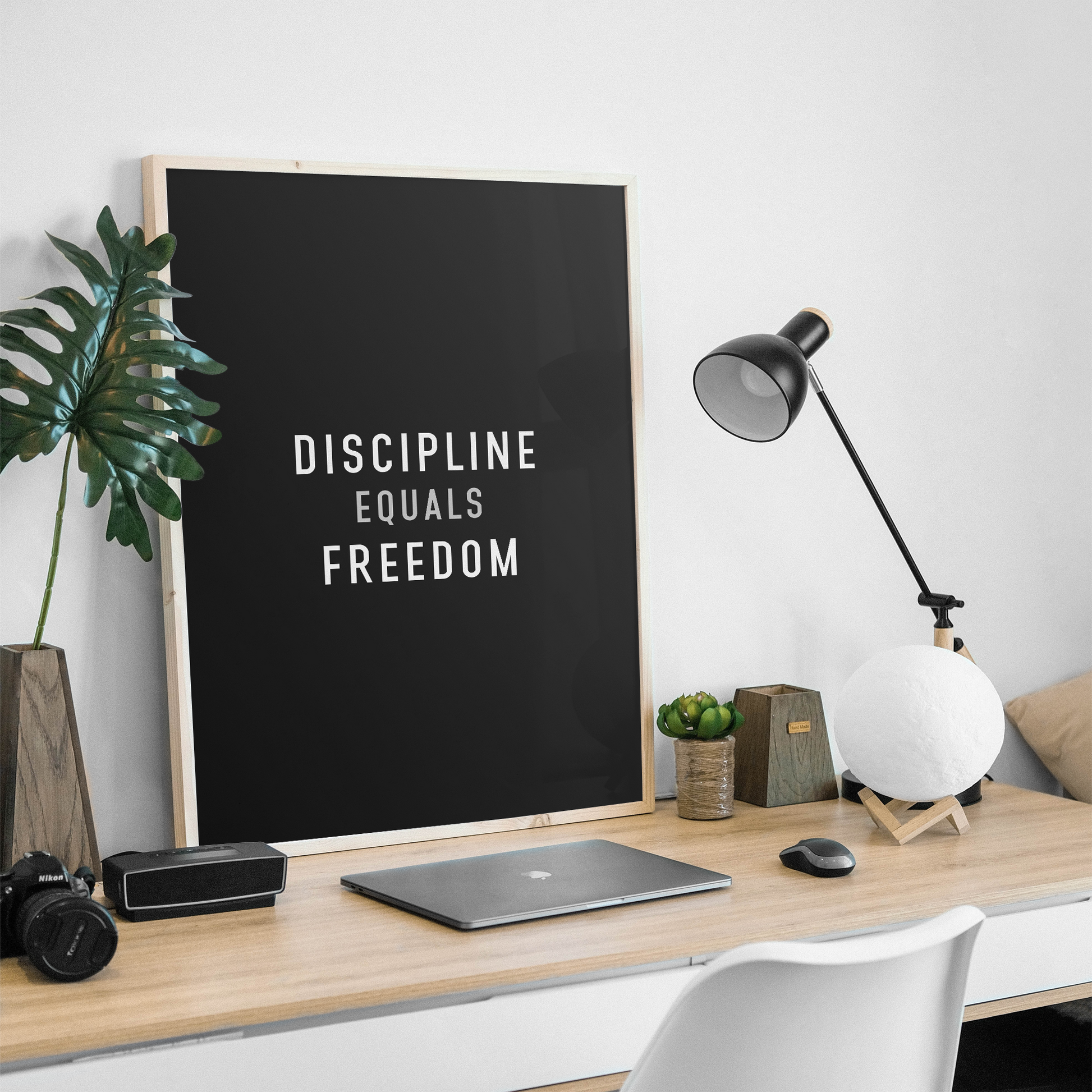 A framed black wall art print with the text 'DISCIPLINE EQUALS FREEDOM' in white, displayed on a light wood desk in a modern home office setting with a laptop, lamp, and plant.